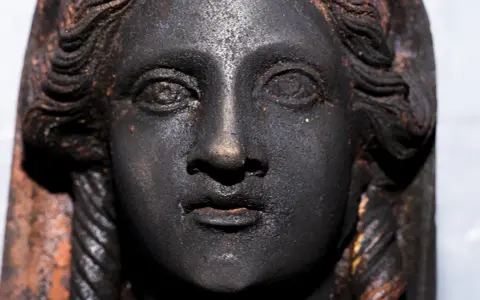 Volto di bronzo scuro, un dettaglio dei bronzi di San Casciano dei Bagni, con capelli a onde e trecce.