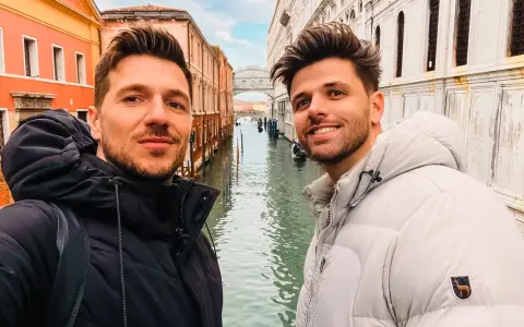 Two men enjoying a romantic gay Venice trip, smiling for a selfie with canals and historic buildings behind them.