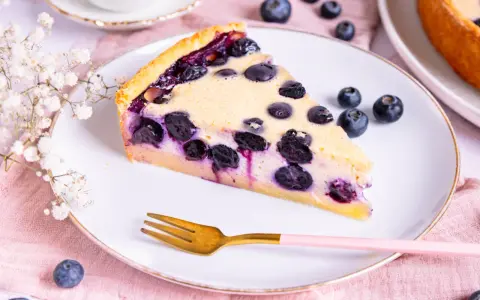 Ein Stück Heidelbeerkuchen Landfrauenart mit saftigen Blaubeeren und cremigem Guss auf einem Teller, daneben eine Gabel und frische Beeren.