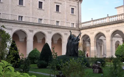 A bronze statue of monks with arms raised stands in a courtyard garden, perhaps commemorating St. Benedict's feast day.