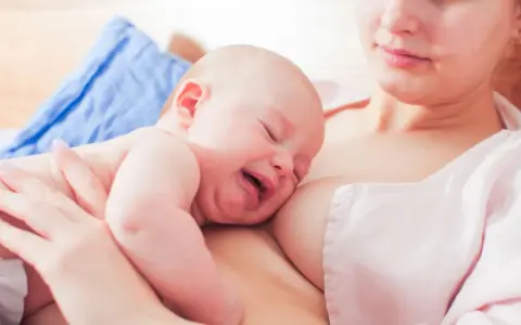 Ein Baby schreit beim Stillen, die Augen geschlossen, das Gesicht verzogen. Die Mutter hält es sanft.