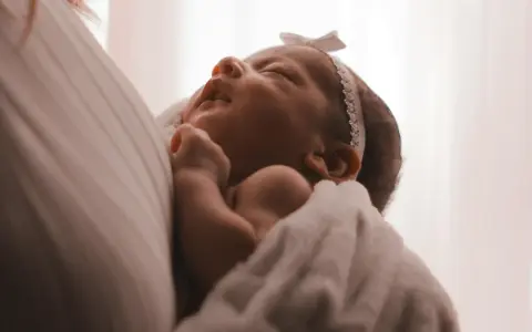 Un neonato avvolto in una coperta bianca, con un fiocco rosa in testa. I suoi piccoli tremori sono quasi impercettibili mentre dorme serenamente.