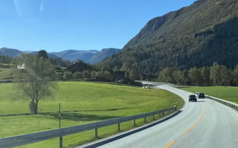 En bil kör på en slingrande väg genom ett grönt landskap med berg och hus. En perfekt start på en semester i Norge.