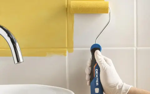 Mano con guante blanco pintando azulejos blancos con rodillo amarillo.