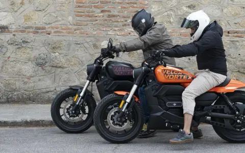 Dos personas en cascos montan dos motos custom electricas, una negra y otra naranja, frente a una pared de piedra.