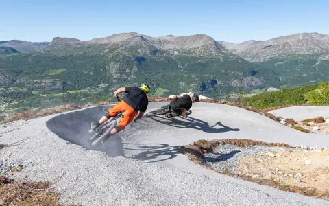 Två cyklister kör utför en grusig bana i Hemsedal sommar. Berg och grönska i bakgrunden.