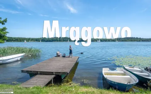 Mrągowo atrakcje: spokojny dzień nad jeziorem, dzieci łowią ryby z pomostu, żaglówki na horyzoncie.
