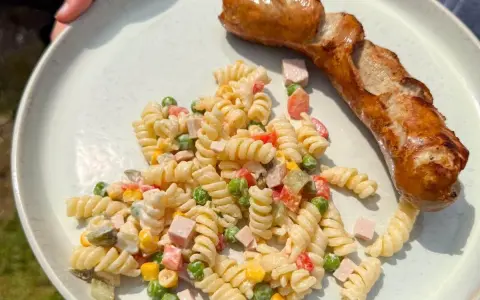 Gegrillte Wurst mit einem cremigen Nudelsalat als Beilage, garniert mit Erbsen, Mais und gewürfeltem Schinken.