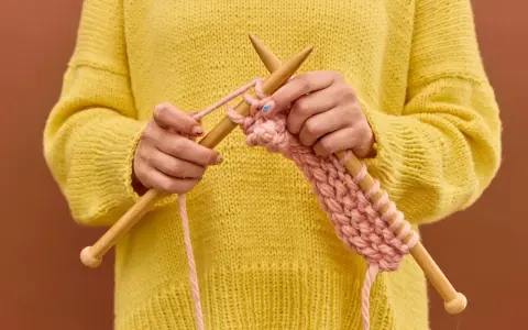 Manos tejiendo un suéter amarillo con hilo rosa, mostrando cómo hacer ochos de 4 puntos.