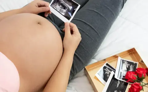 Brzuch w 12 tygodniu ciąży, z USG w dłoniach. Obok pudełko z kolejnymi zdjęciami i różami.