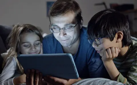 Padre y dos hijos ven una tablet, sonriendo. Podrían estar aprendiendo sobre los riesgos de las redes sociales o disfrutando de contenido familiar.
