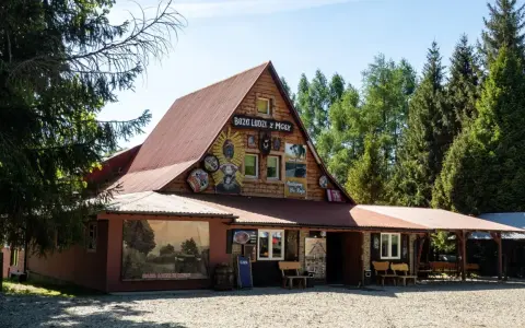 Drewniany budynek z napisem "Baza Ludzi z Mgły" i malowidłami. Obok duży mural z krajobrazem i napisem "Baza Ludzi z Mgły".