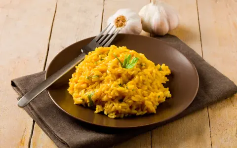 Un invitante risotto zafferano in polvere, guarnito con prezzemolo fresco, su un piatto marrone, con aglio e forchetta sullo sfondo.