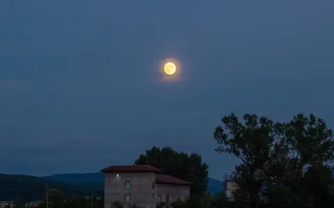 La luna di luglio illumina un paesaggio serale con colline, alberi e un edificio.