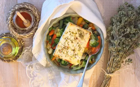 Feta al forno con verdure miste, condita con erbe aromatiche. Accanto, olio, spezie e un mazzetto di origano secco.