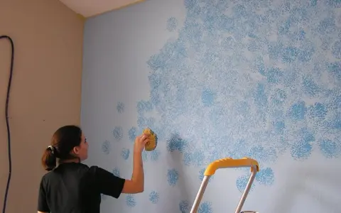 Mujer aplica pintura que disimula imperfecciones en pared con esponja, creando un patrón de nubes azules.