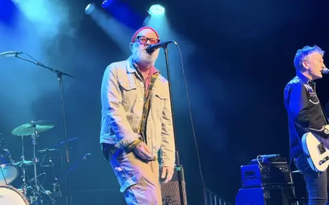 Le chanteur principal du rem groupe, avec une barbe blanche et un bonnet rouge, chante sur scène. Un guitariste est à droite.