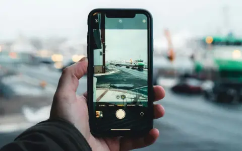 Dłoń trzyma aparat na telefon, rejestrując zimowy widok portu z zielonym statkiem i samochodami na drodze.