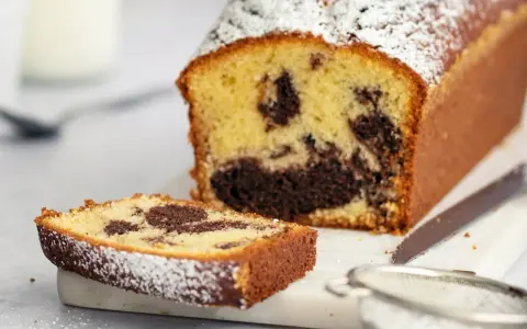 Ein saftiger Marmorkuchen in Kastenform, bestäubt mit Puderzucker. Eine Scheibe liegt daneben, bereit zum Genuss.