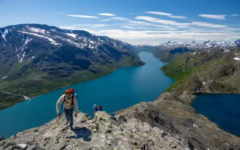 Vandrare klättrar på en bergskam med en turkos sjö nedanför. Besseggen svårighetsgrad är utmanande men belönande.