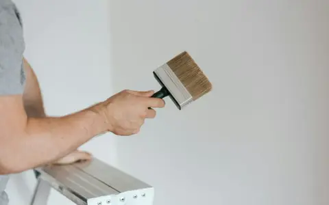 Person på en stege håller en bred pensel redo att måla huset vitt.