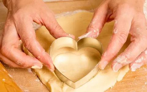 Manos cortan masa con un cortador en forma de corazón, parte de una receta de galletas.