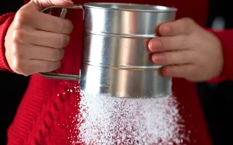 Hände halten einen Siebbecher, aus dem feiner Zucker rieselt. So einfach lässt sich puderzucker selber machen für süße Kreationen.