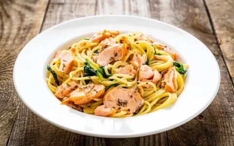 Makaron spaghetti z łososiem i szpinakiem na białym talerzu, na drewnianym tle.