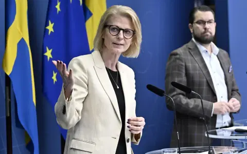 Kvinna med glasögon talar vid presskonferens med svenska och EU-flaggor bakom. Viktig information om folkräkning Sverige.