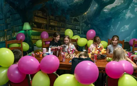 Dzieci świętują urodziny w pokoju zagadek. Kolorowe balony i owocowy tort tworzą radosną atmosferę.