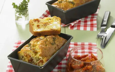 Cake tomates séchées feta, un délice salé aux herbes fraîches. Une tranche est posée sur un cake encore dans son moule.