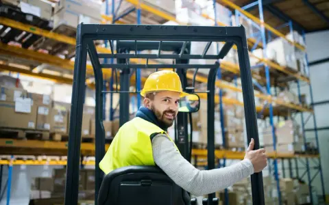 Uśmiechnięty operator wózka widłowego w żółtym kasku i kamizelce odblaskowej, z kciukiem w górze. Zastanawiasz się, ile zarabia operator wózka widłowego?