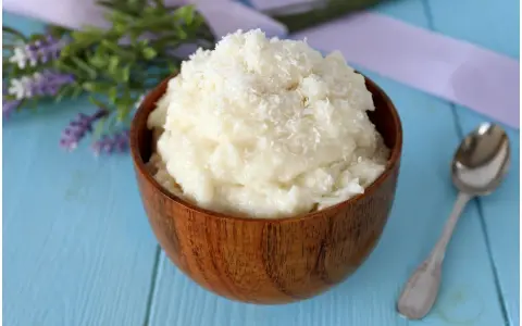 Una ciotola di legno piena di cremosa crema al cocco, guarnita con cocco grattugiato. Accanto, un cucchiaino d'argento e fiori di lavanda.