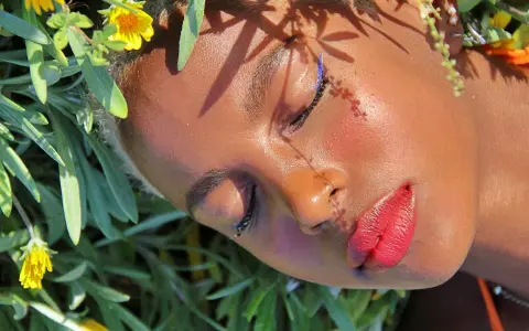 Un volto immerso tra fiori, con un trucco festival audace: eyeliner blu elettrico e labbra rosse vibranti.