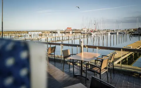 Ein Tisch mit Stühlen auf einer Holzterrasse mit Blick auf einen Yachthafen. Segelboote liegen im Wasser, die Sonne wirft lange Schatten. Ein Möwe fliegt über den Hafen. Damp 2000.
