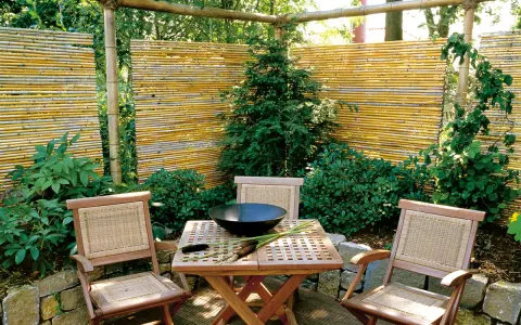Ein lauschiger Sitzplatz mit einem selbstgemachten Zaun aus Bambus. Holzmöbel und üppige Pflanzen schaffen eine natürliche Atmosphäre.