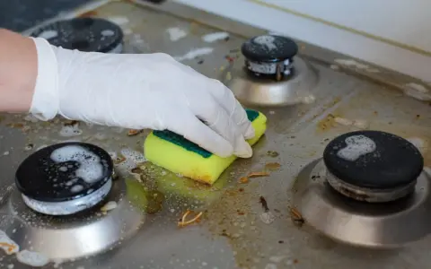 Ręka w rękawiczce czyści zabrudzoną kuchenkę gazową gąbką. Poznaj sposoby, jak wyczyścić kuchenkę, by znów lśniła.