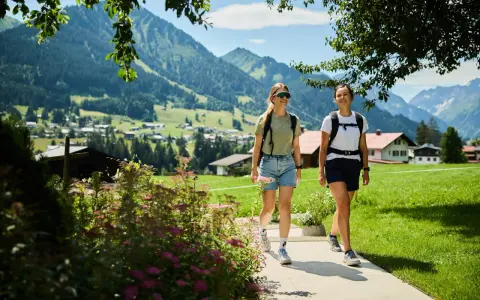 Zwei Frauen wandern durch das Kleinwalsertal mit Blick auf Berge und Häuser. Ein kleiner See ist nicht zu sehen.