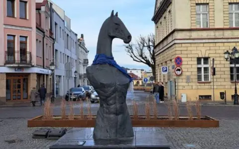 Pomnik konia w Koninie, z niebieską szarfą na szyi, stoi na brukowanym placu, otoczony kamienicami.