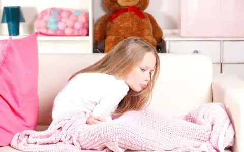 Bambina di 12 anni con mal di stomaco, rannicchiata sul divano sotto una coperta rosa.