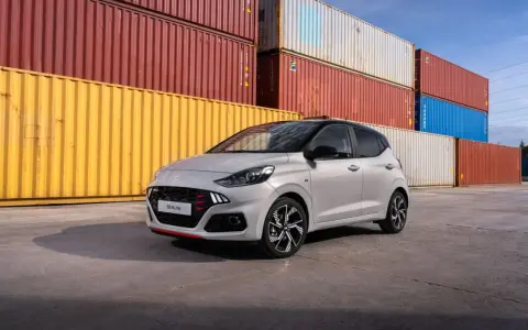 Szary Hyundai i10 N Line z czerwonymi akcentami na grillu i zderzaku, zaparkowany przed stosem kolorowych kontenerów. Sprawdź atrakcyjną hyundai i10 cena!