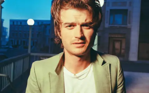 Joe Keery w jasnej marynarce, z naszyjnikiem, na tle nocnego miasta.