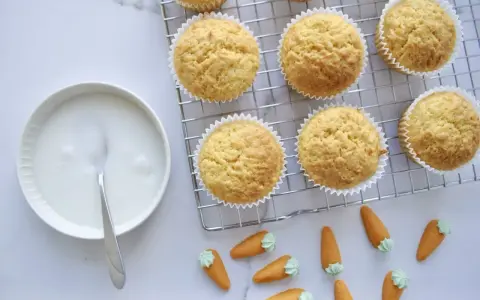 Goldbraune Karottenmuffins auf einem Gitter, daneben eine Schüssel mit Zuckerguss und süße Marzipankarotten. Perfekt für ein Karottenmuffins Rezept!