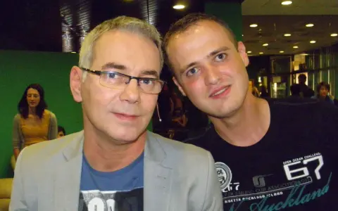 Tomasz Kammel i młody fan pozują do zdjęcia. Tomasz Kammel w szarym garniturze i niebieskiej koszulce, a obok niego młody mężczyzna w czarnej koszulce z napisem "Auckland".