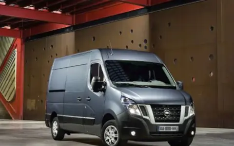 Szary Nissan NV400 stoi przed brązową, perforowaną ścianą w industrialnym wnętrzu.