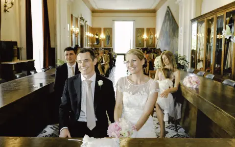 Uśmiechnięty świadek na ślubie, obok panny młodej, w eleganckim garniturze. W tle goście i dekoracje sali.