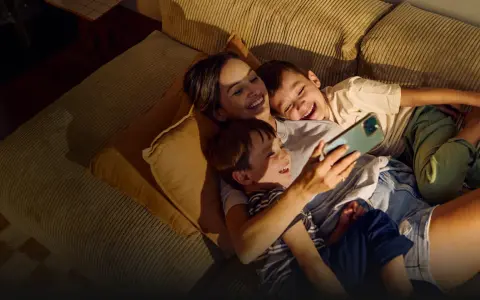 A mother and two sons laugh while looking at a smartphone on a couch.