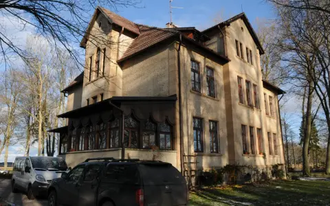 Dawny pałac Poraj, z charakterystyczną werandą i drzewami w tle, otoczony zimowym krajobrazem.