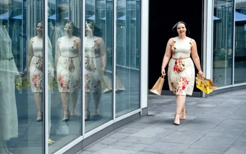 Kobieta w kwiecistej sukience, z szerokimi biodrami, idzie z torbami na zakupy, odbijając się w lustrzanej fasadzie.