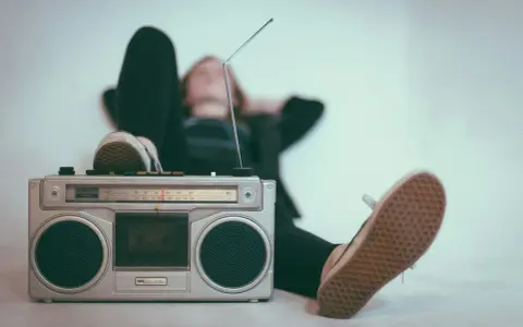 Osoba relaksuje się przy radiomagnetofonie, słuchając muzyki. To pokazuje, jak łatwo dotrzeć do odbiorców, co jest jedną z **zalet reklamy radiowej**.
